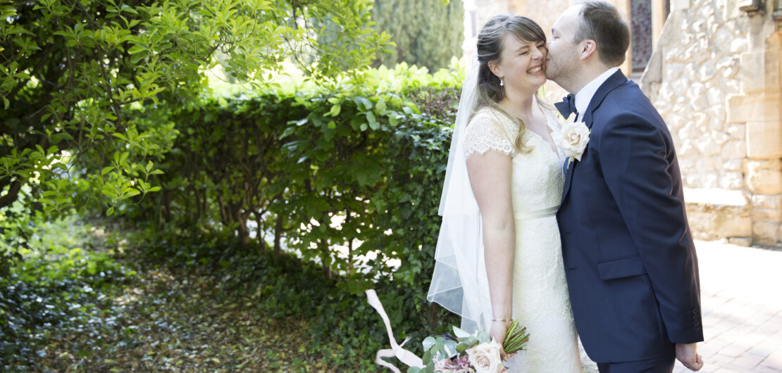 Bride laughing being kissed by groom in wedding portrait outside of St Stephen's Church in Tonbridge, Kent