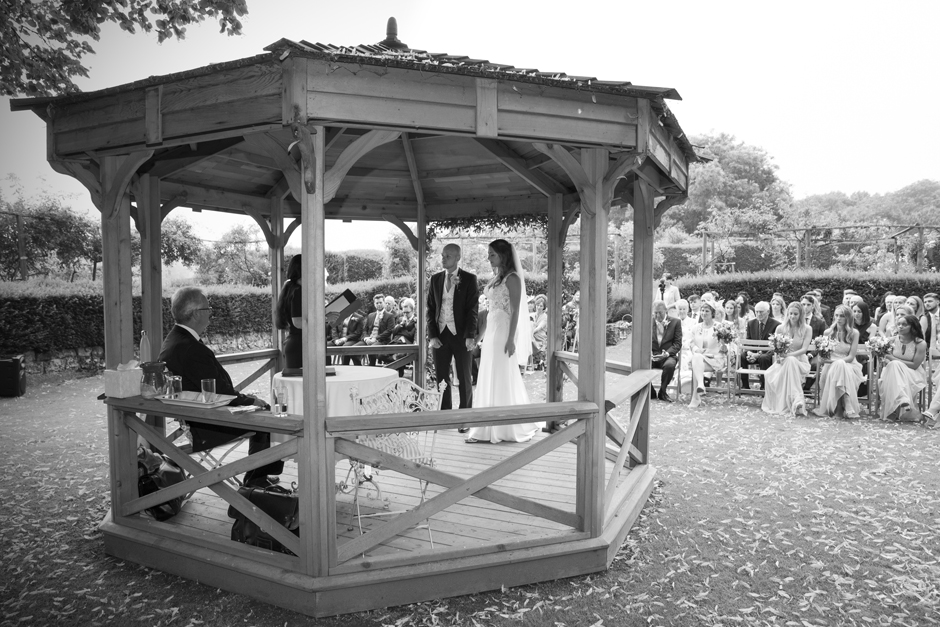 Front of wooden gazebo with guests behind and bride and groom standing under. Captured at Nettlestead Place by Victoria Green Photography.
