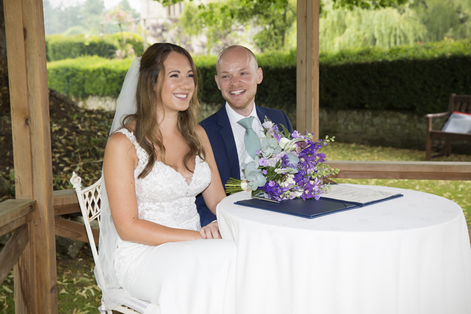 Bride and groom laughing at table signing the register at outside wedding ceremony at Nettlestead Place in Kent. Captured by Kent wedding photographer, Victoria Green.