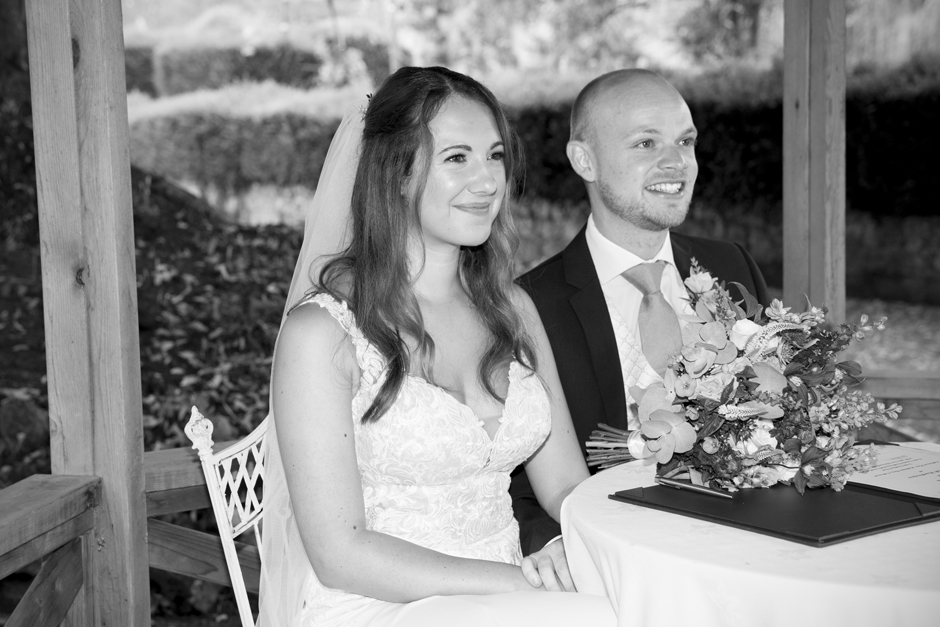 Bride and groom smiling sitting at table where they have just signed the register. Captured at outside wedding ceremony at Nettlestead Place in Maidstone, Kent. Captured by photographer Victoria Green.