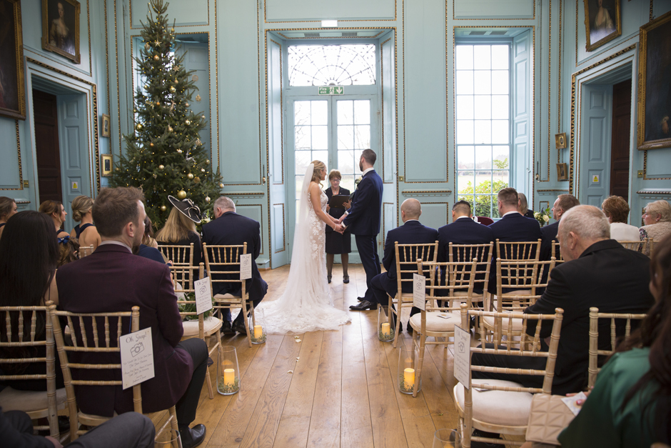Wide shot of back of wedding ceremony room with guests onlooking at bride and groom hold hands during their vows at Bradbourne House in East Malling, Kent.
