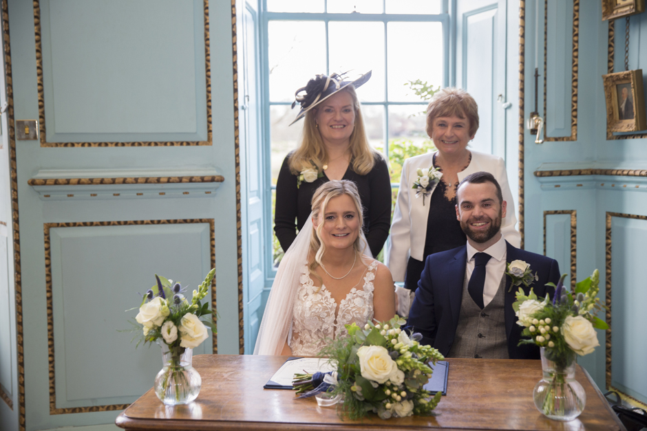 Bride and Groom sitting at table after signing the register with their female witnesses behind them during wedding ceremony at Bradbourne House in East Malling, Kent.