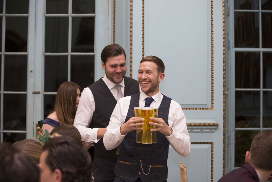 Male wedding guests laughing holding glasses full of beer during wedding breakfast at Bradbourne House in East Malling, Kent.