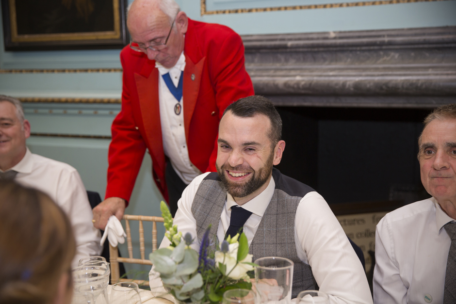 Groom laughing seated during wedding breakfast with toastmaster in background at Bradbourne House in East Malling, Kent.