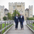 Grooms holding hands walking over bridge at Hever Castle in Kent. Wedding Photography by Victoria Green.