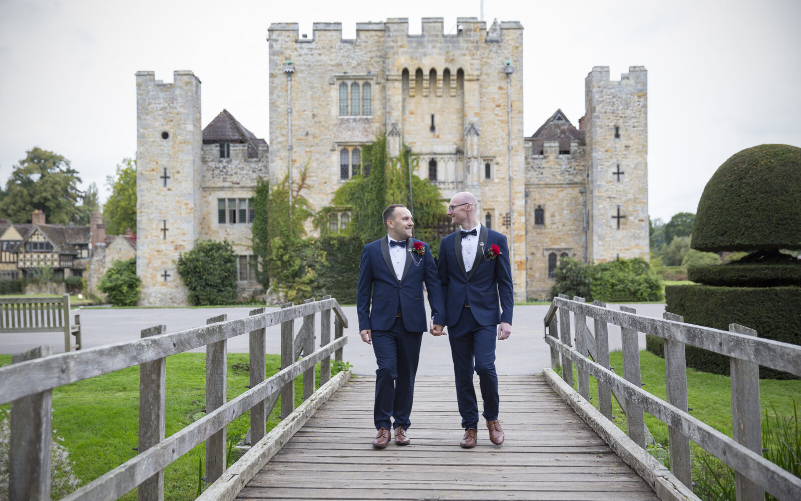 Grooms holding hands walking over bridge at Hever Castle in Kent. Wedding Photography by Victoria Green.