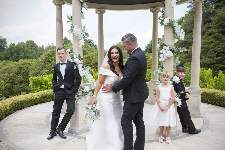 Bride and Groom laughing together with their children posing by the pillars of The Spa Temple at The Spa Hotel, Tunbridge Wells in Kent. Captured by Kent wedding photographer Victoria Green.